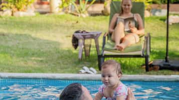 Una piscina con una persona y un niño en el agua, una silla de descanso y una zona cubierta con plantas.