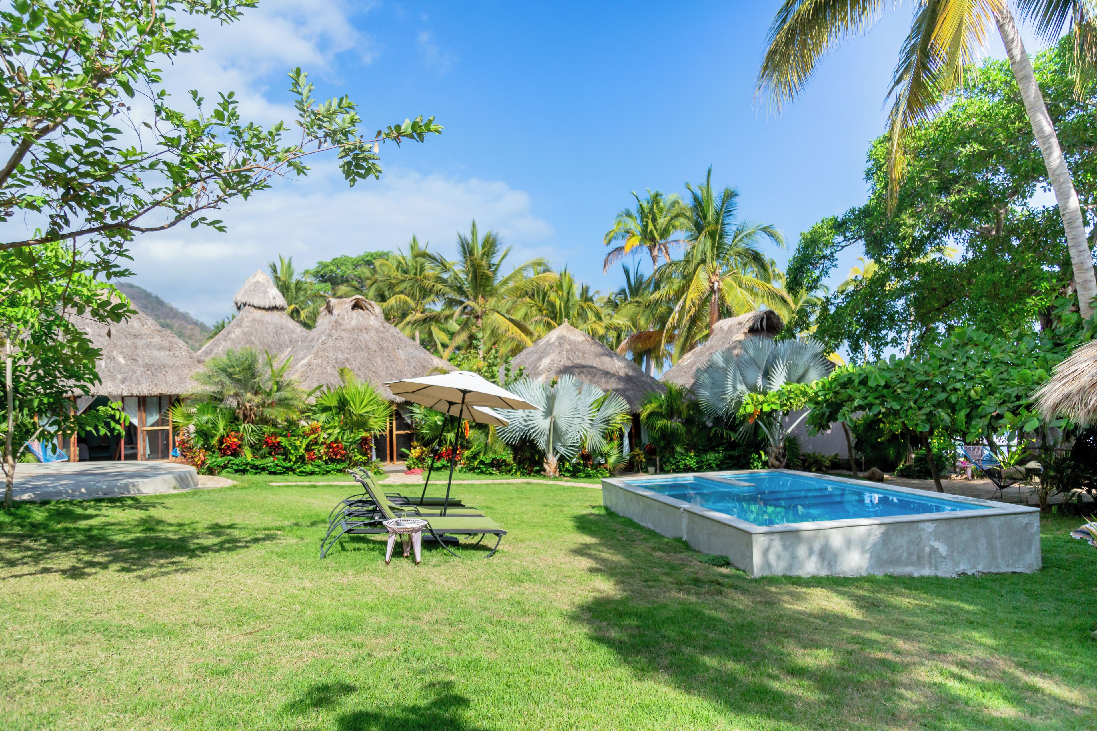 Zona de piscina con sillas de descanso y sombrillas, rodeada de vegetación tropical y construcciones con techo de paja.