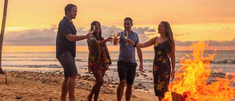 Un grupo de personas alrededor de una fogata en la playa, con una puesta de sol al fondo.