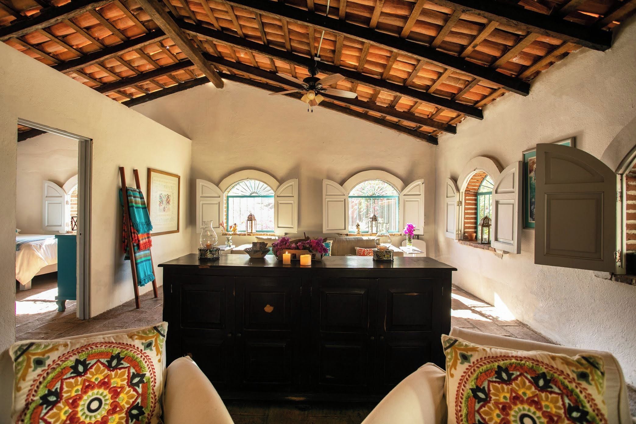 A room with a wooden ceiling, a central island with a vase and candles, and a view through arched windows.