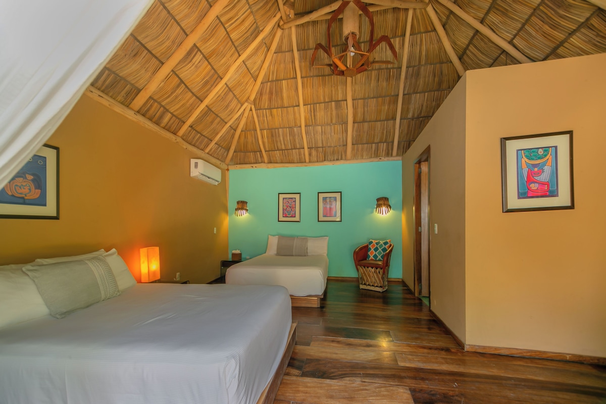 A room with a thatched roof, a bed with white bedding, a chair with a colorful cushion, and wall-mounted lights.
