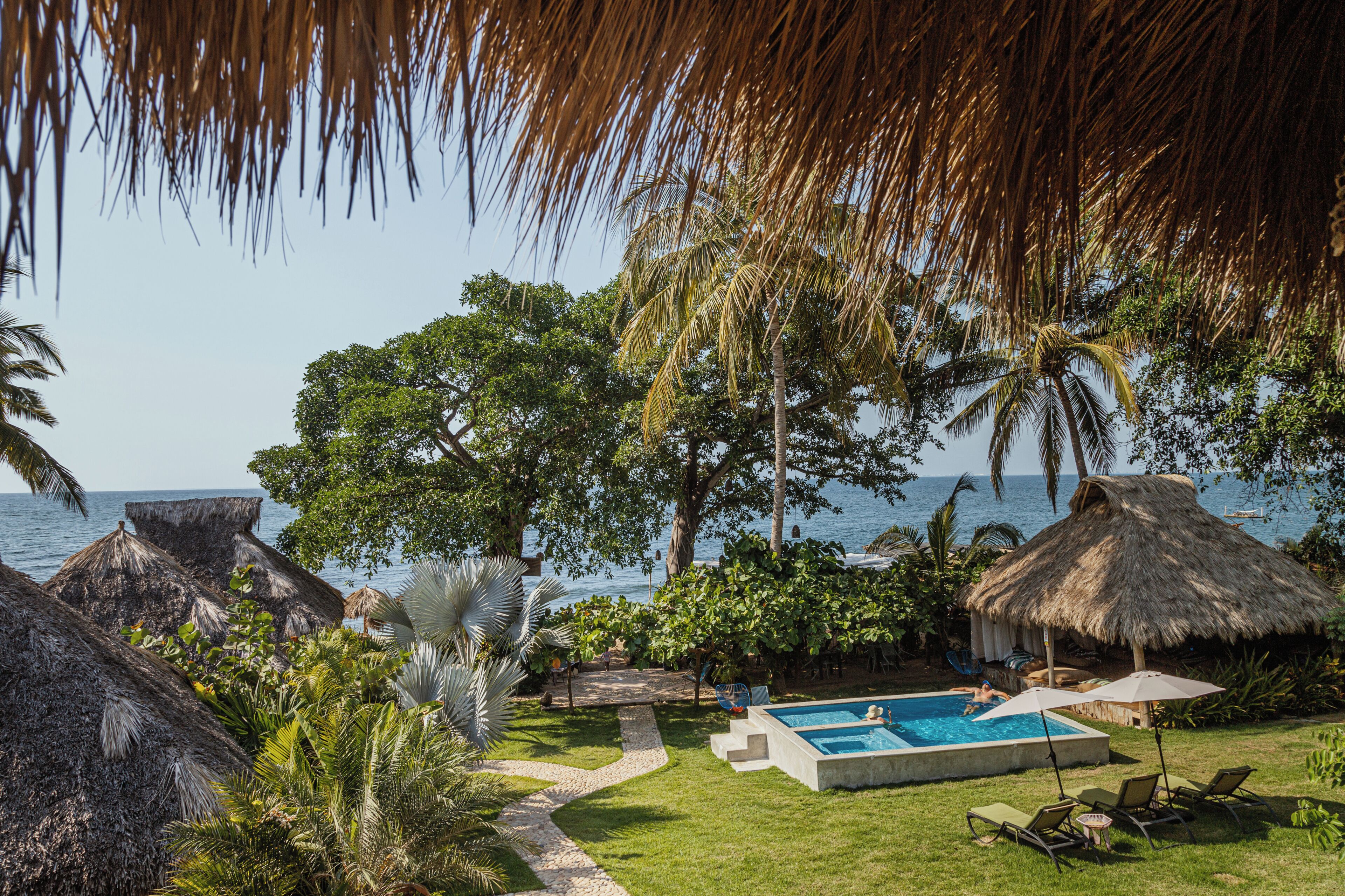 Zona de piscina con vistas despejadas al océano, rodeada de exuberante vegetación y construcciones de techo de paja.