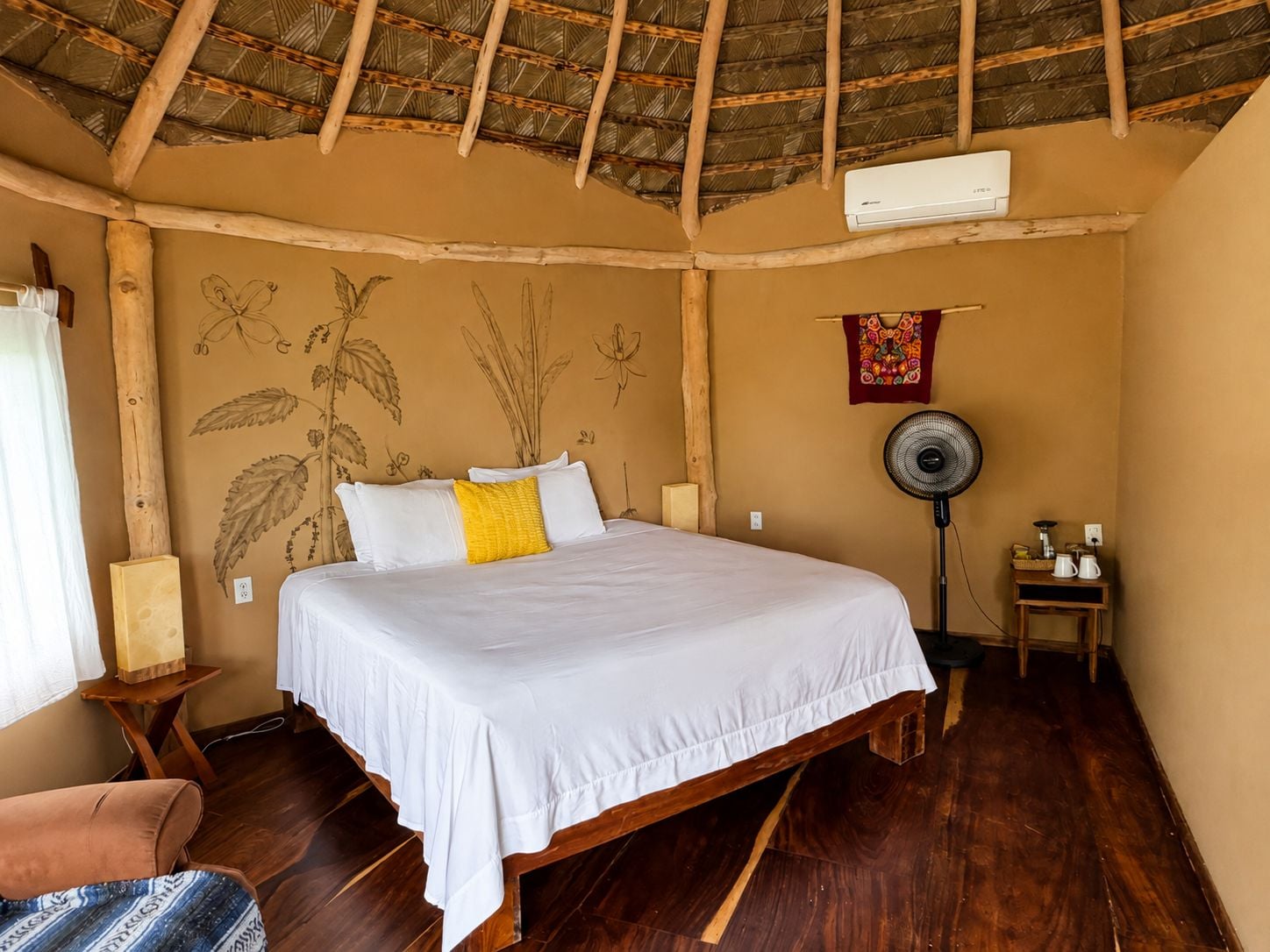 A room with a bed, a fan, a table with a kettle, and a wall with botanical illustrations.
