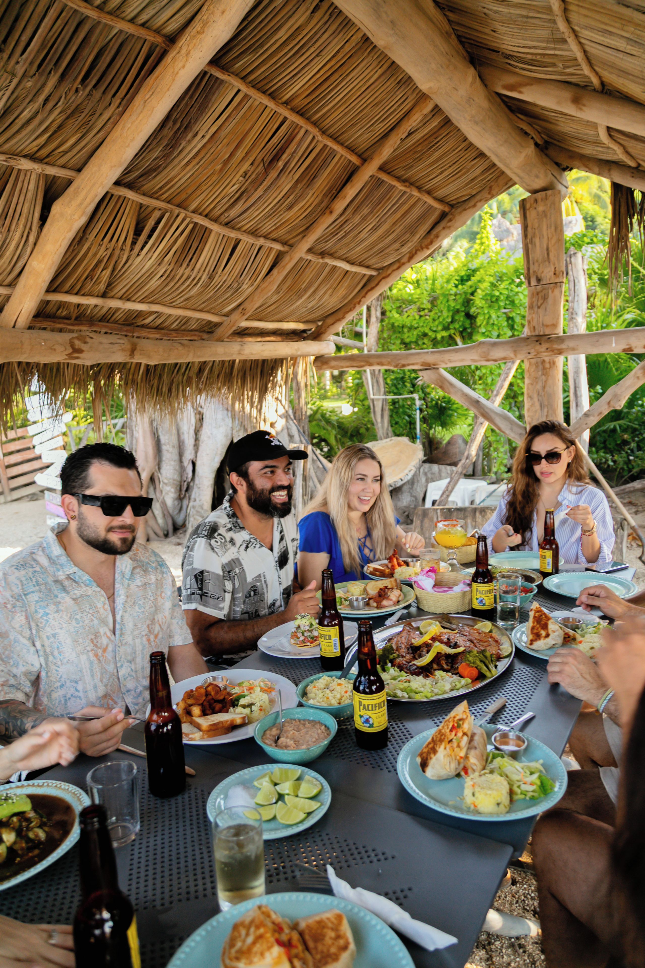 Un grupo de personas cenando en una mesa al aire libre bajo un tejado de paja, rodeado de follaje.