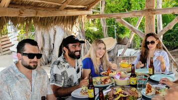 Un grupo de personas cenando en una mesa al aire libre bajo un tejado de paja, rodeado de follaje.