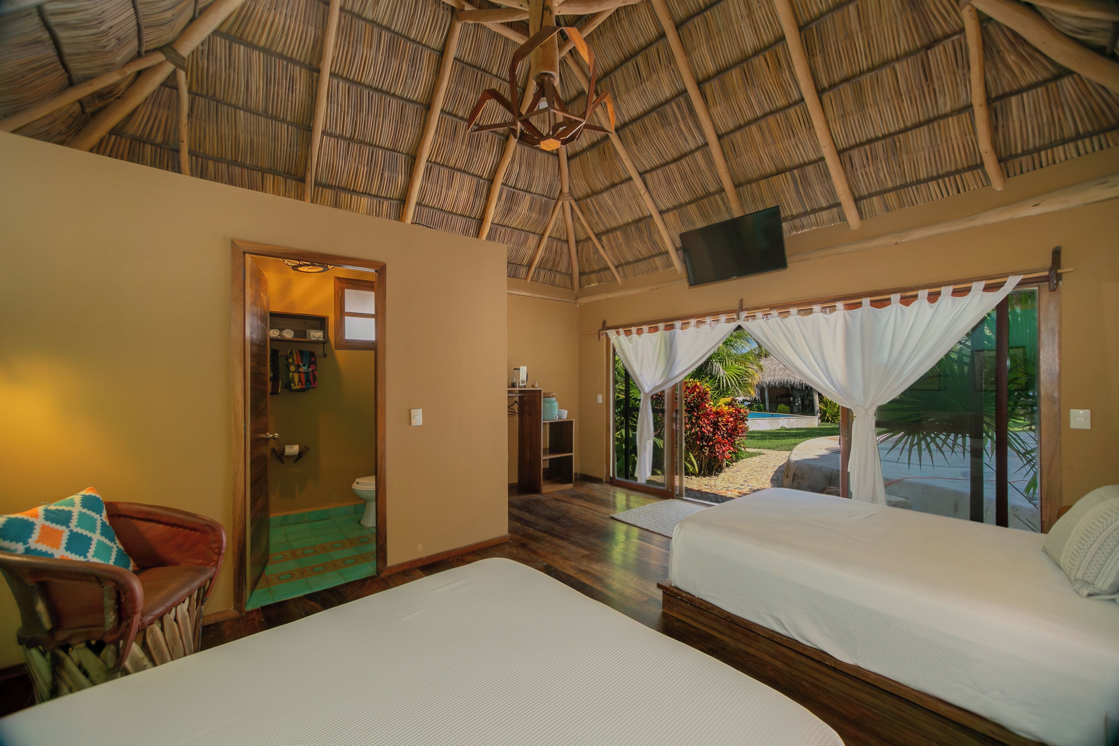 A room with a thatched roof, two beds, a wooden chair, a television, and a view of a garden and pool outside.