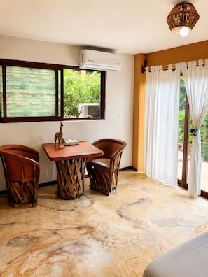 A room with a table and two chairs, an air conditioning unit, a window with a view of greenery, and a curtain. - Amaca Beach Retreat - Puerto Vallarta South Shore (Quimixto)