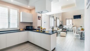 Una cocina moderna con armarios blancos, encimera negra y una isla central. Hay un comedor con mesa y sillas blancas, y una sala de estar con sofá y mesa de centro.