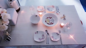Ein ordentlich gedeckter Esstisch mit tellerförmiger Blumenmusterdeko, ein Teller mit Kirschtorte-Motiv und eine Kerze.