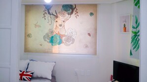 Ein Schlafzimmer mit einem Bett, einem Wandbild, einer Deckenleuchte und einem Fenster mit Blick auf Grünflächen.