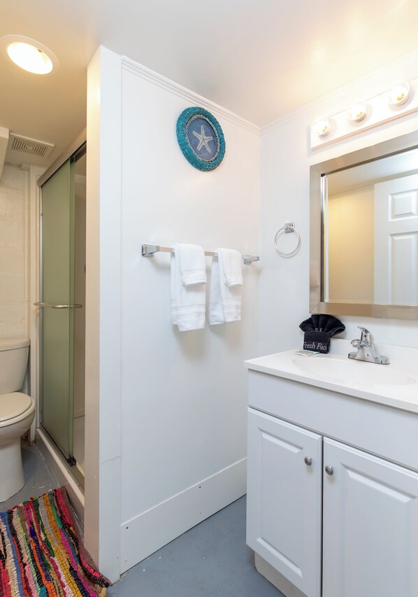 A bathroom with a white toilet, a shower with a glass door, a white sink with a mirror, and a colorful rug on the floor.
