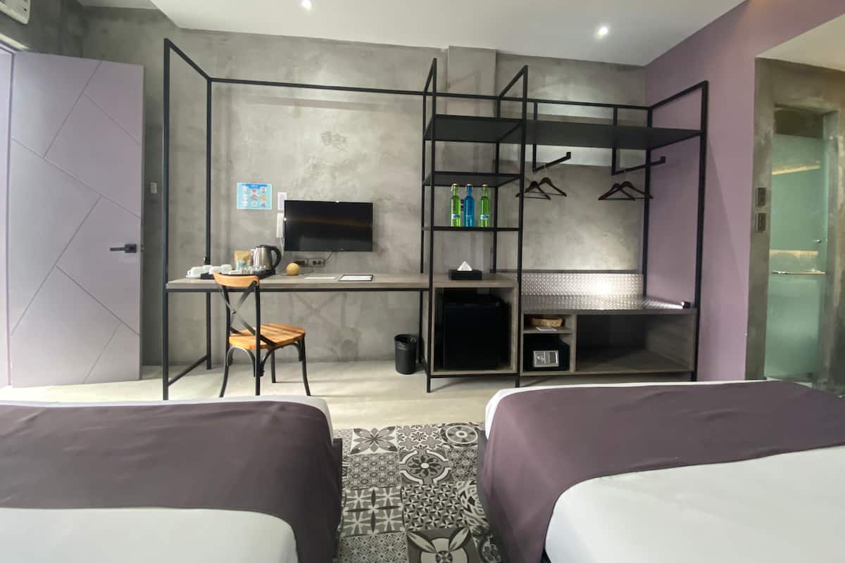 A modern hotel room with two beds, a desk with a computer, a TV, and a seating area with a chair and shelves.