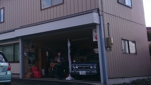 Un bâtiment à deux étages avec une porte de garage ouverte, laissant apercevoir une voiture à l’intérieur. On peut voir une enseigne avec des caractères japonais sur la porte de garage.