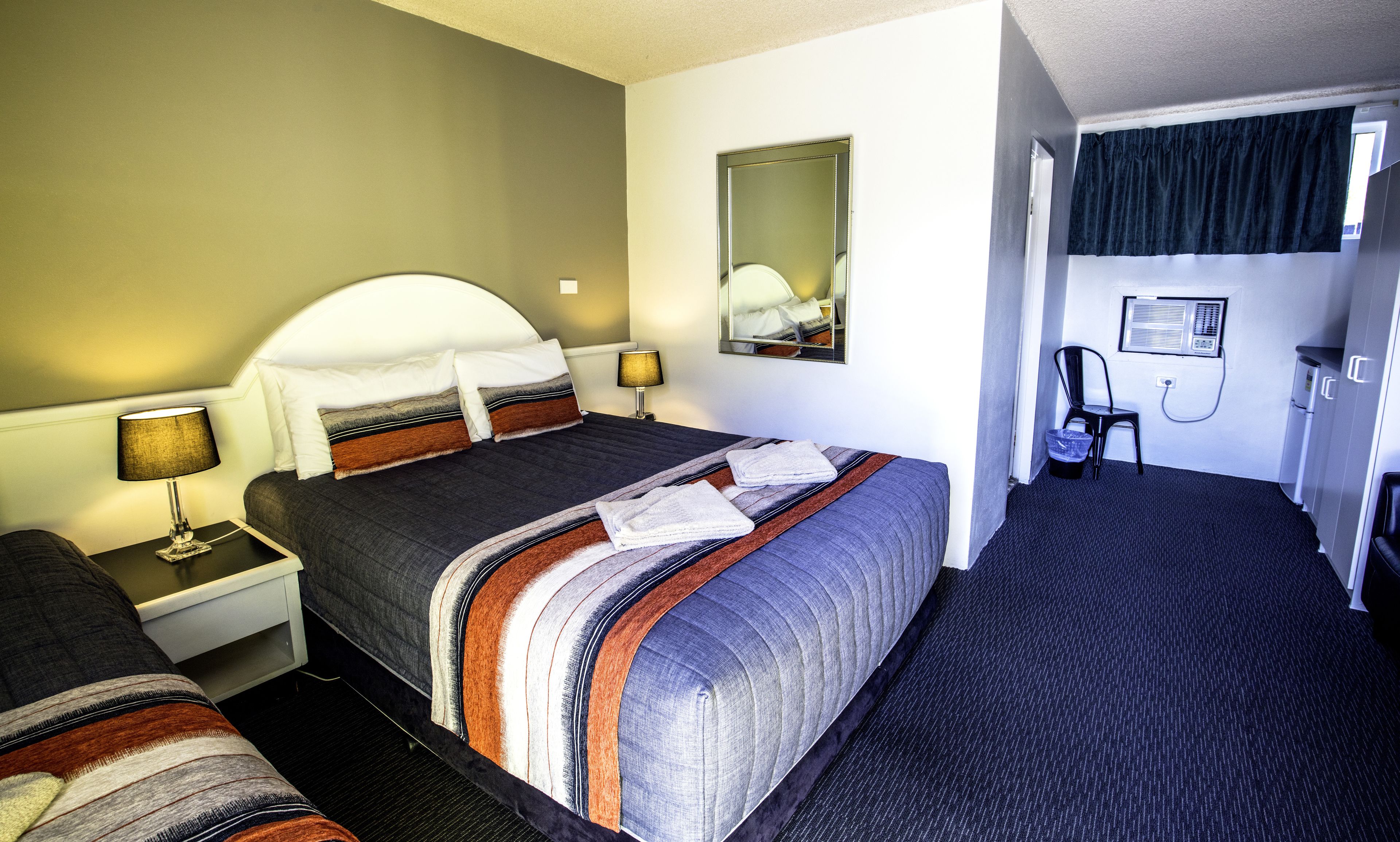 A hotel room with a bed, a nightstand, a lamp, a mirror, a chair, a desk, and a wardrobe.