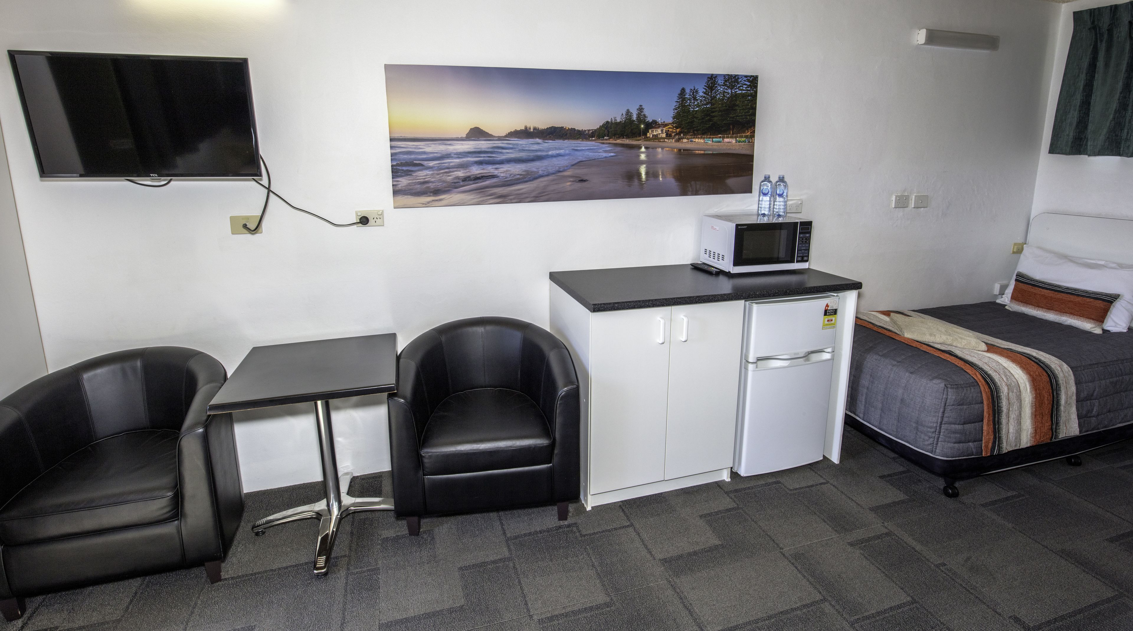 A compact hotel room with a kitchenette, a bed, a desk with two chairs, a flat-screen TV, and a painting of a beach scene.