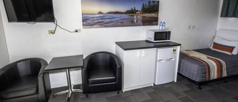 A compact hotel room with a kitchenette, a bed, a desk with two chairs, a flat-screen TV, and a painting of a beach scene.