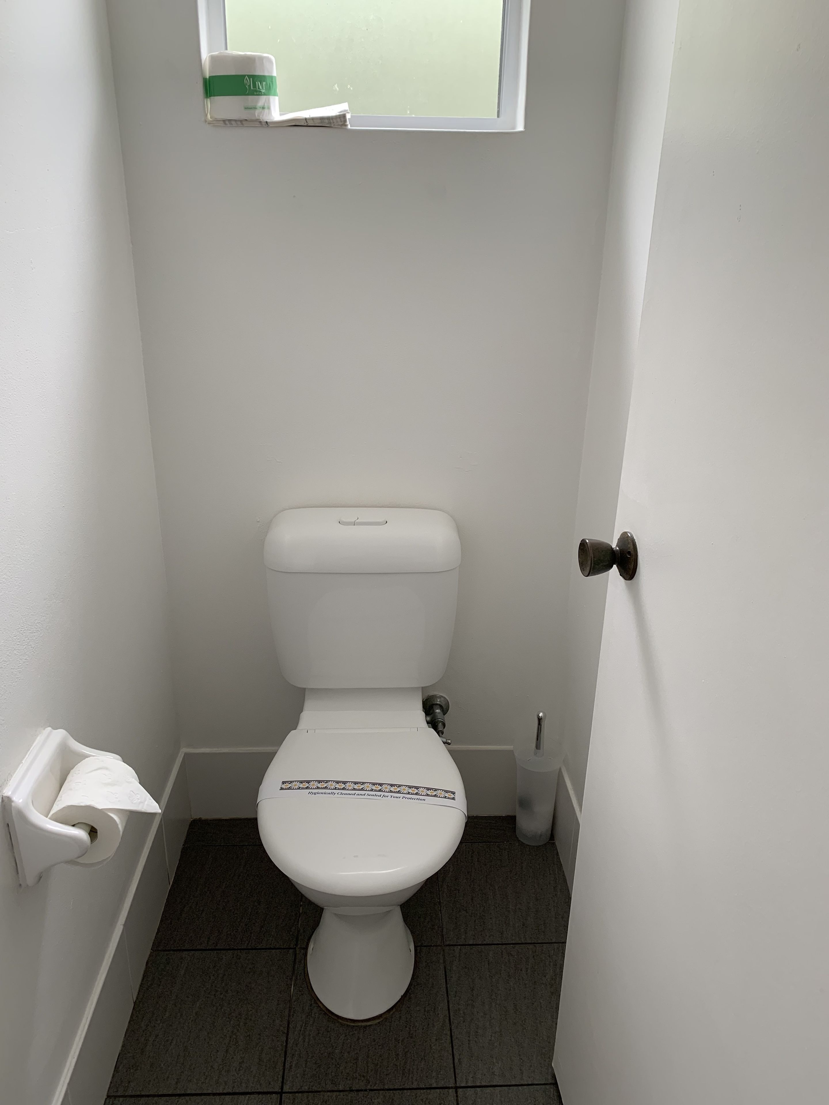 A small, white bathroom with a toilet, a window above it, and a towel dispenser.