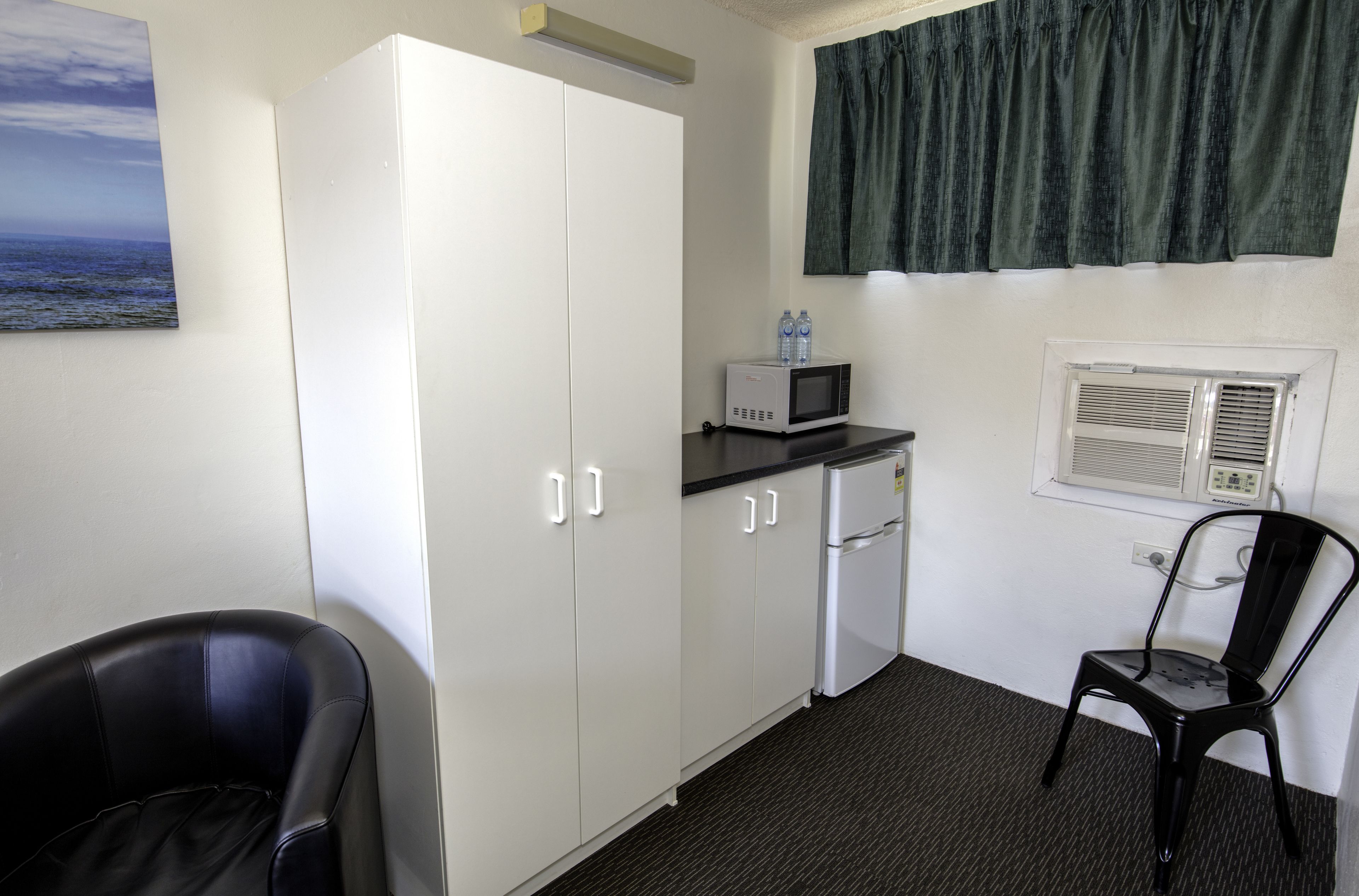 Ein kleines Zimmer mit einer Küchenzeile, einem schwarzen Stuhl, einem weißen Kleiderschrank und einem Fenster mit Vorhang.