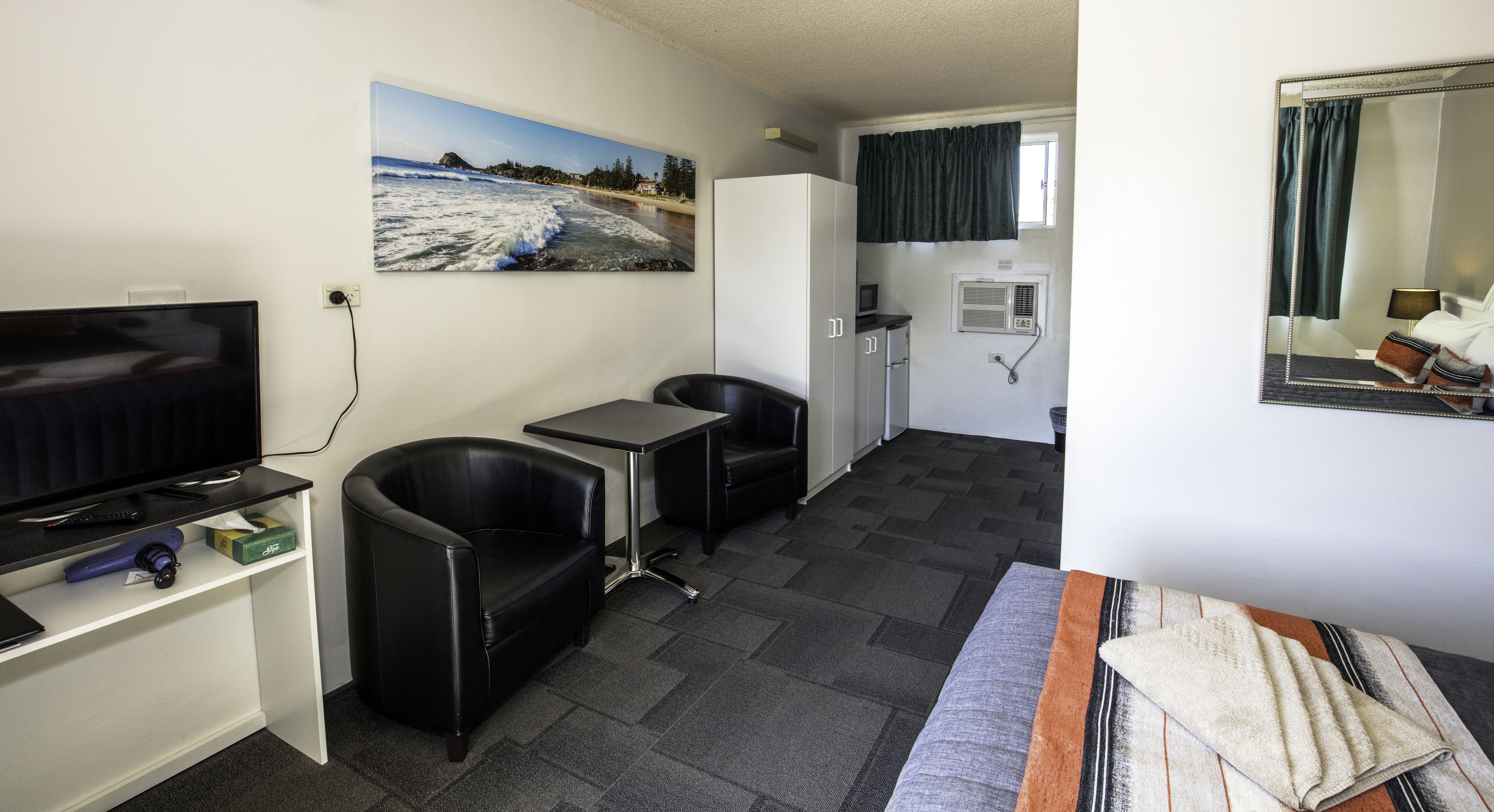Ett kompakt hotellrum med en tv, ett litet bord, två stolar, en soffa, en tavla som föreställer en strand, ett fönster med gardiner och en spegel.