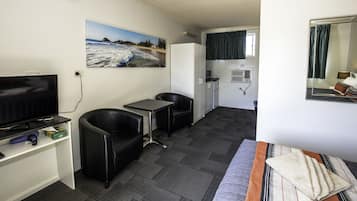 Ett kompakt hotellrum med en tv, ett litet bord, två stolar, en soffa, en tavla som föreställer en strand, ett fönster med gardiner och en spegel.