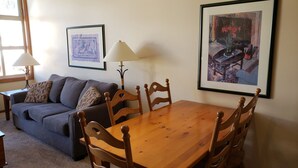 A living room with a wooden dining table, four chairs, a grey sofa, two lamps, and two framed artworks on the walls. - Cozy, ski-in/out, condo in the heart of Sun Peaks (Thompson-Nicola P)