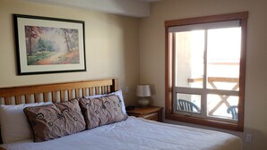 A bedroom with a large bed, a nightstand, a lamp, a framed painting, and a window with a view. - Cozy, ski-in/out, condo in the heart of Sun Peaks (Thompson-Nicola P)