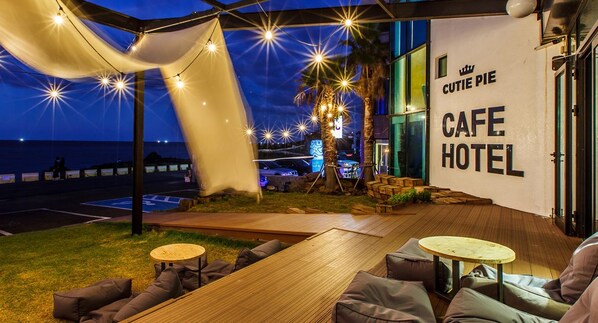 A cozy outdoor seating area with wooden tables and cushioned chairs, a palm tree, and a view of the sea. - Cutie Pie Hotel (Jeju City)