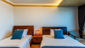 Quarto de hotel com duas camas, uma mesinha de cabeceira com abajur e uma cadeira.