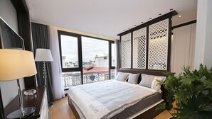 Habitación de hotel con una cama grande, un escritorio con lámpara, una planta en maceta y vistas a la ciudad a través de amplias ventanas.
