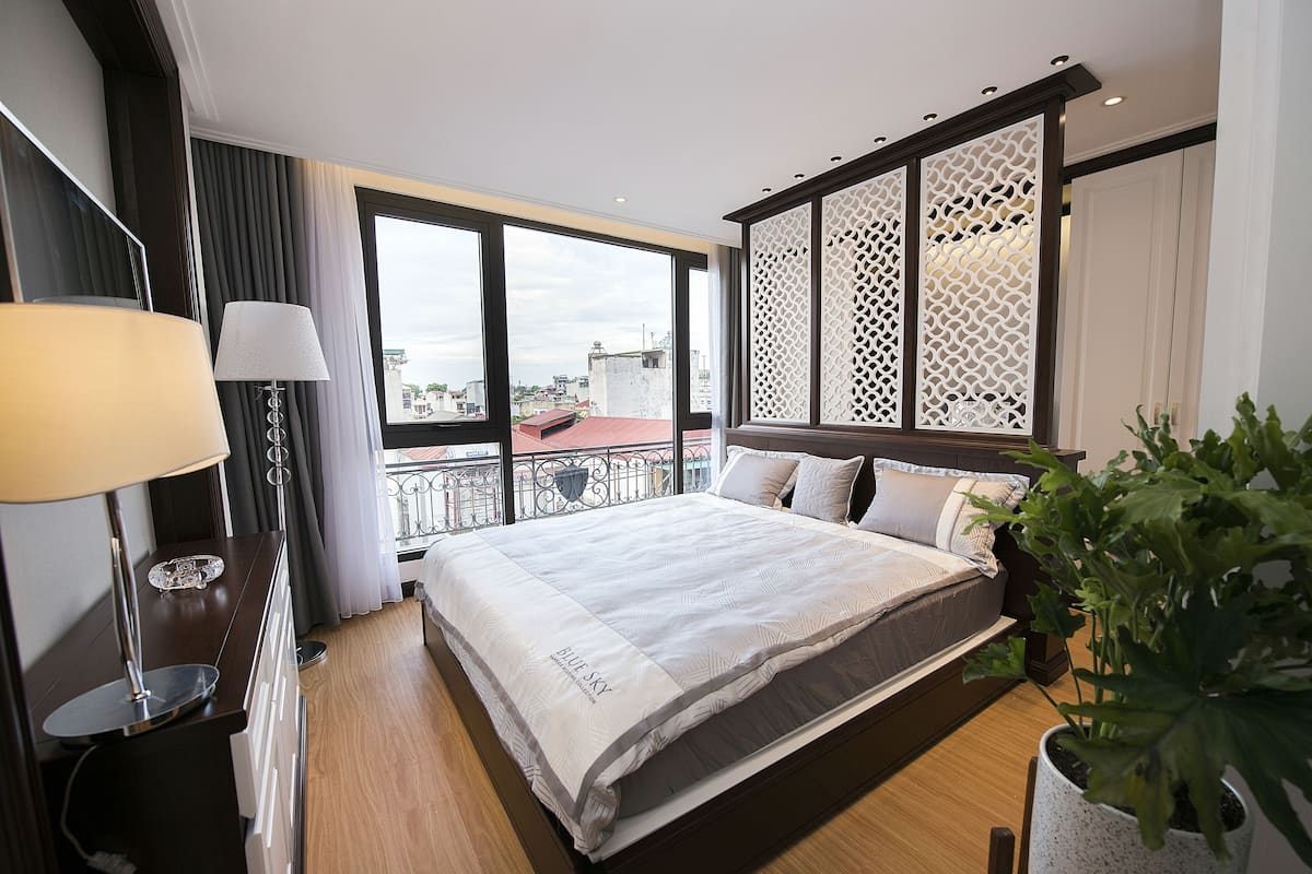 A hotel room with a large bed, a desk with a lamp, a potted plant, and a view of the city through large windows.