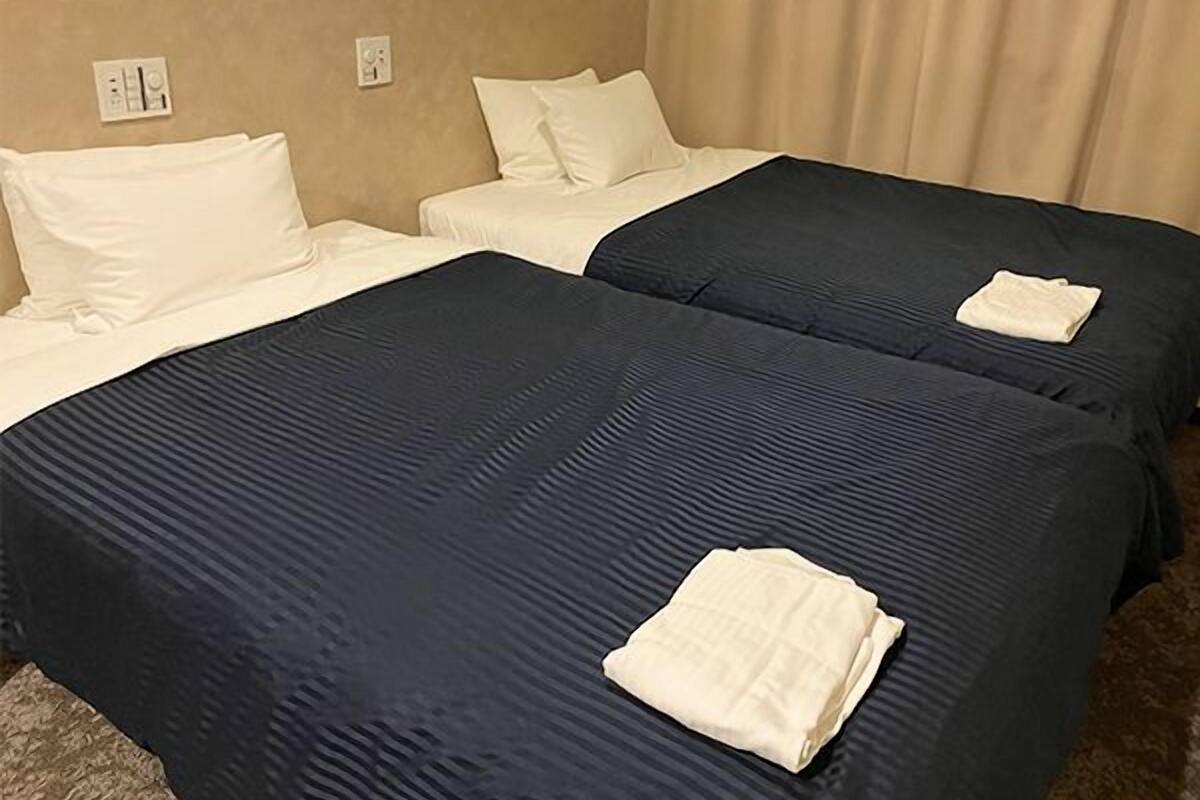 A hotel room with two beds, each with a dark blue bedspread and white pillows. There is a folded towel on one of the beds.
