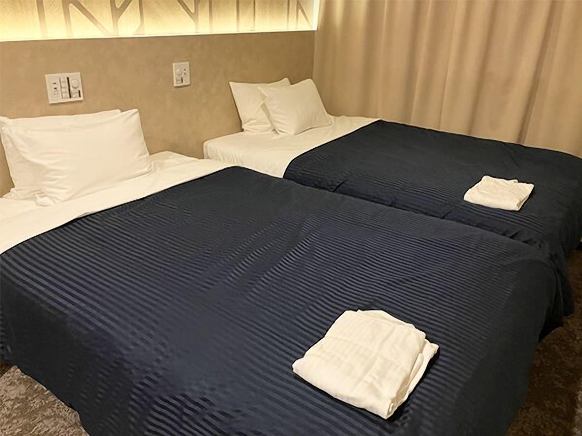A hotel room with two beds, each with a dark blue bedspread and white pillows. There are two wall sockets visible.