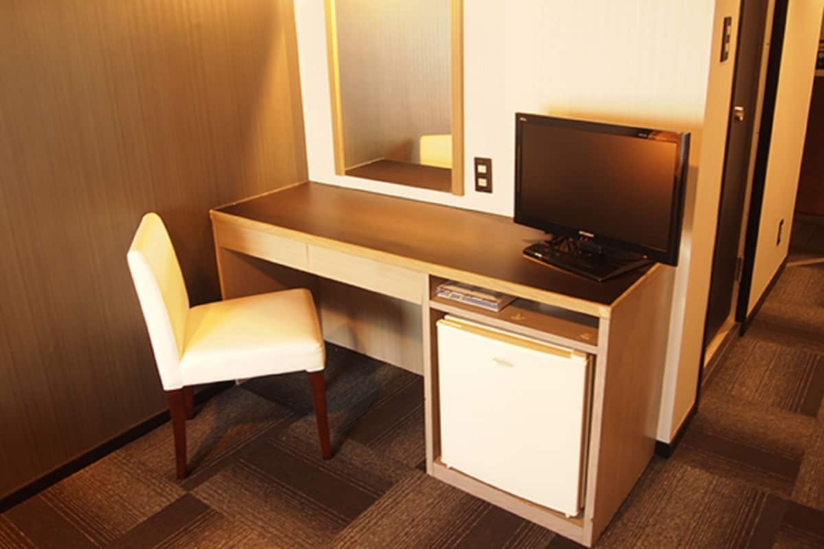 A compact hotel room with a small desk, a chair, a television, and a mini-fridge.
