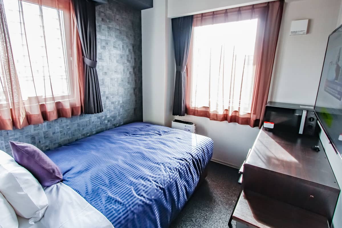A hotel room with a bed, a desk, a chair, and two windows with curtains.