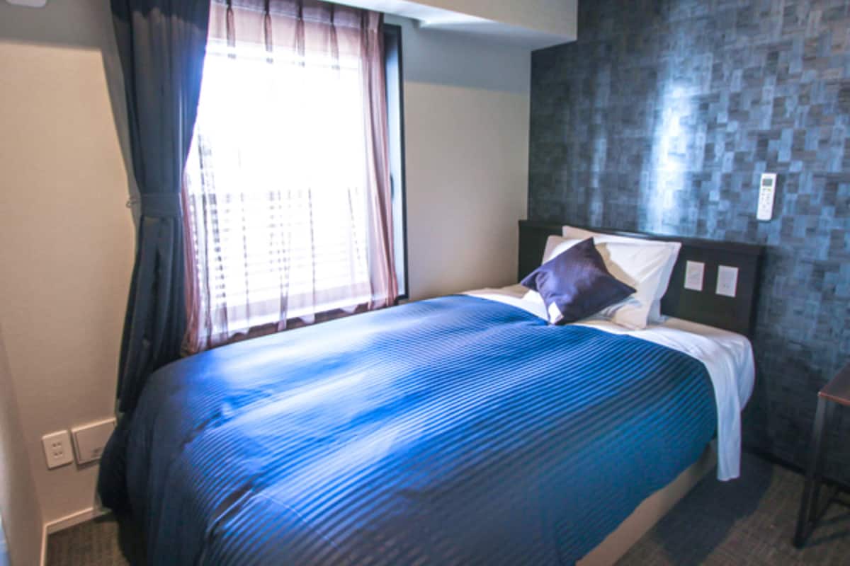 A hotel room with a bed, a window with curtains, a wall with a textured pattern, and a bedside table.