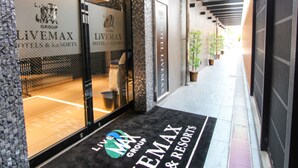 Entrada de un hotel con fachada de vidrio, felpudo negro con el nombre del hotel y un pasillo con plantas en macetas.