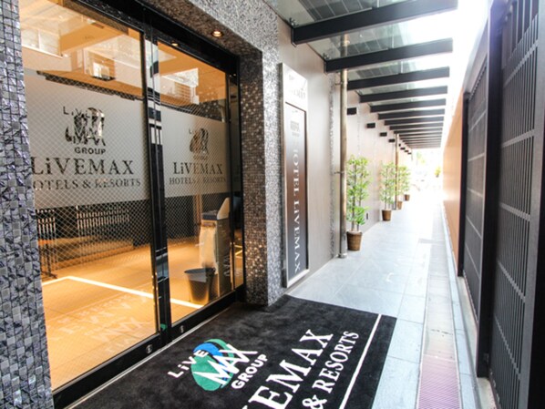 A hotel entrance with a glass facade, a black doormat with the hotel's name, and a corridor with potted plants.