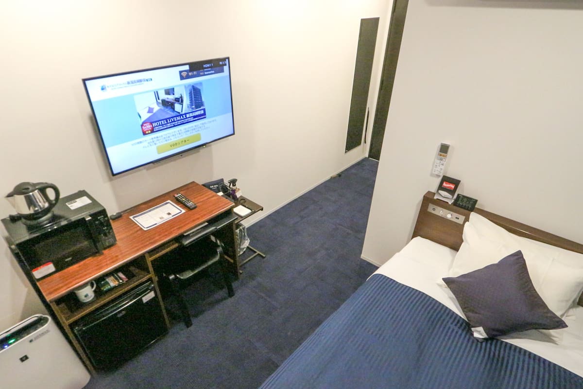 Ein Hotelzimmer mit Bett, Schreibtisch mit Computer, Kaffeemaschine und Fernseher, der eine Hotelanzeige zeigt.
