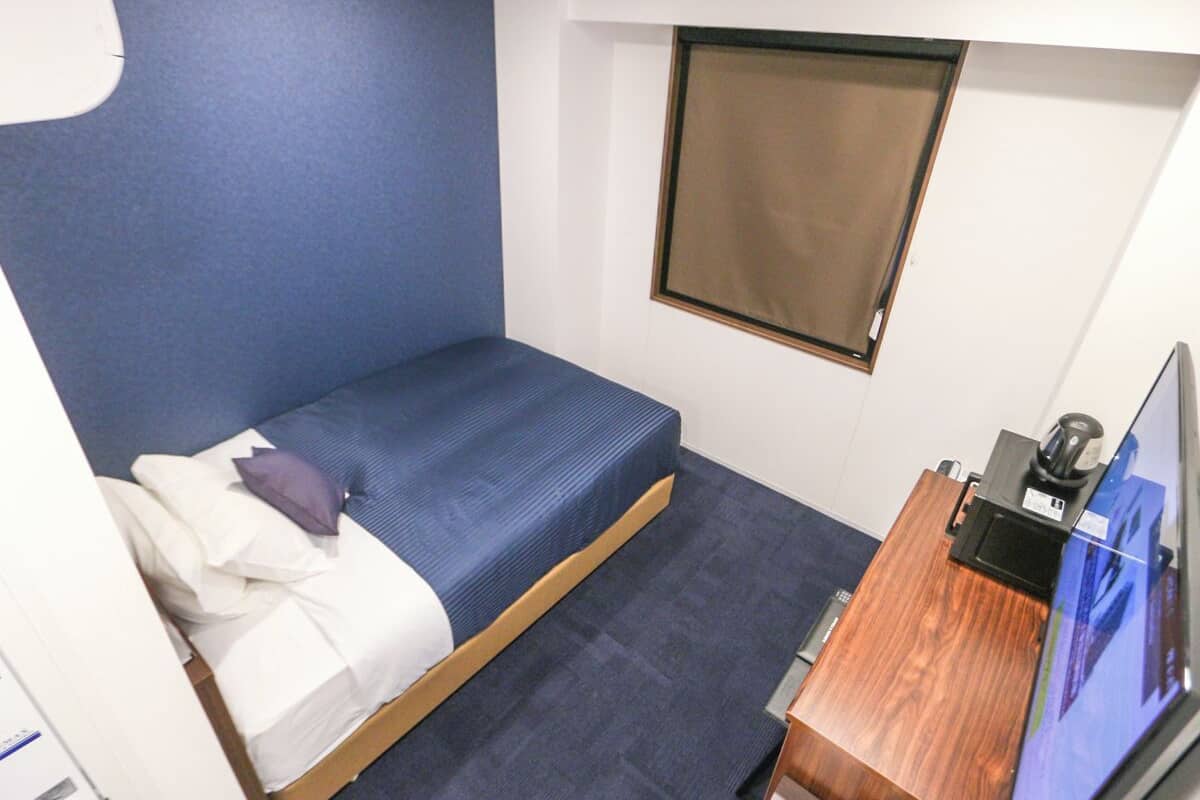 Ein kleines Zimmer mit einem Einzelbett, einem blauen Kopfteil, einem Holztisch mit Computer und einem Fenster mit Jalousien.
