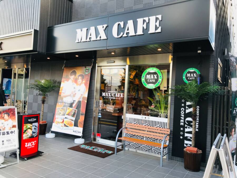 The exterior of a cafe with a prominent 'MAX CAFE' sign, a bench outside, and a potted plant to the right.