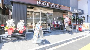Ein Hotel namens 'HOTEL LIVEMAX' mit einem angrenzenden 'MAX CAFE'. Vorgelagerte Blumenarrangements, ein Schild und ein bepflasterter Gehweg.