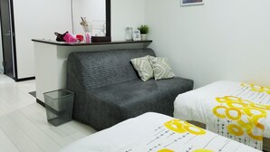Et moderne hotelværelse med en grå sofa, to senge med gul og hvidt sengetøj, en lille hylde med toiletartikler, et spejl og et indrammet billede på væggen.