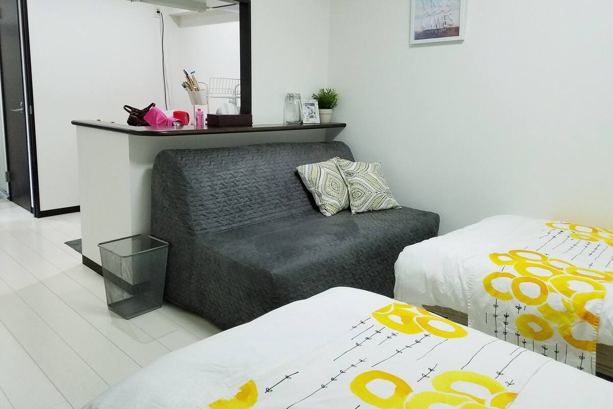 Une chambre d’hôtel moderne avec un canapé gris, deux lits avec une literie jaune et blanche, une petite étagère avec des articles de toilette, un miroir et un tableau encadré au mur.