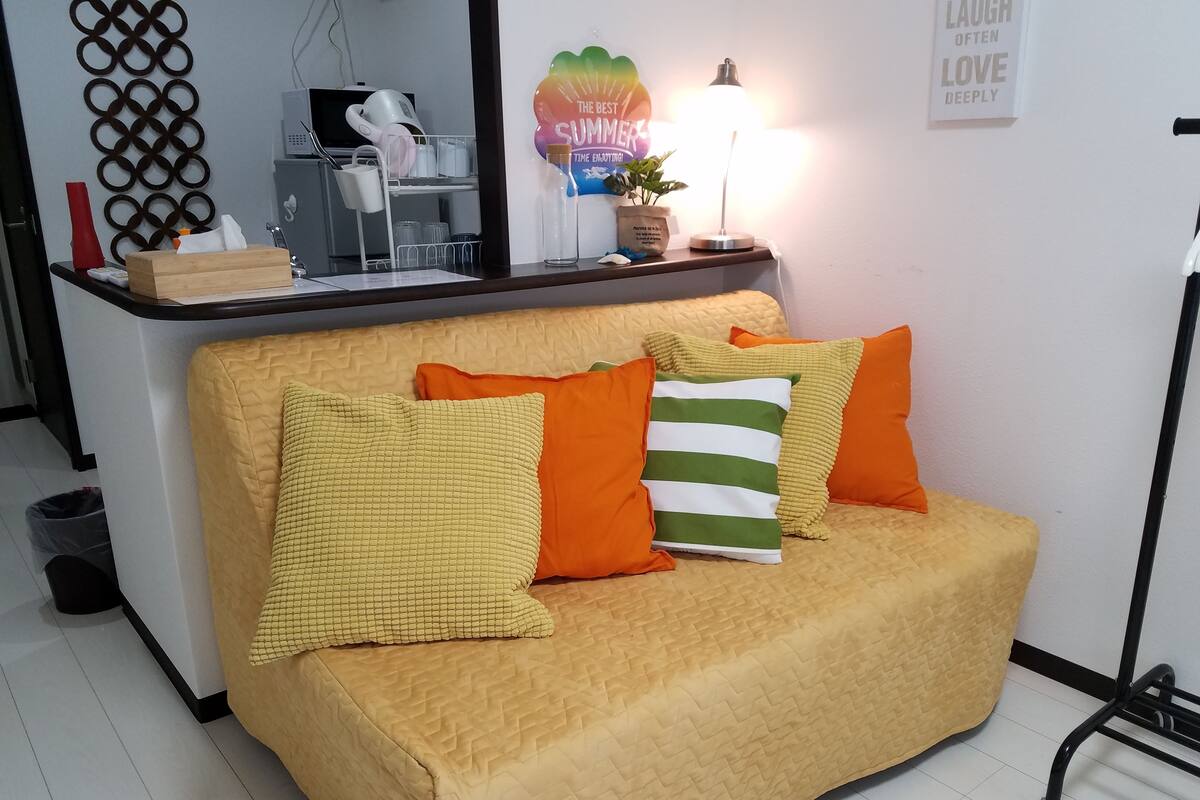 Un canapé jaune agrémenté de coussins orange et verts, une petite table avec une lampe et un élément décoratif mural.