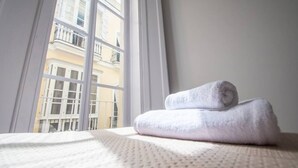 A neatly made bed with a rolled white towel, a window with a view of a building with balconies. - Apartamento Musa Catalina (Cádiz)