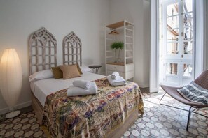 Un lit bien fait, recouvert d’une couvre-lit à motifs floraux, une lampe de chevet et une table de chevet blanche agrémentée d’une plante.