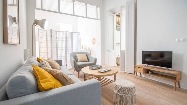 A modern living room with a grey sofa, a round coffee table, a small round ottoman, a TV on a wooden stand, and a large window with a view. - Apartamento Musa Catalina (Cádiz)