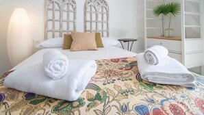A neatly made bed with floral-patterned bedding, two decorative headboards, a bedside table, and a shelf with a plant. - Apartamento Musa Catalina (Cádiz)