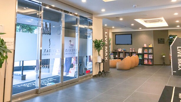 A modern hotel lobby with a glass entrance, seating area with orange chairs, a 'Live Max' sign, and a wall-mounted TV. - HOTEL LiVEMAX Kyoto Kamogawamae (Kyoto)