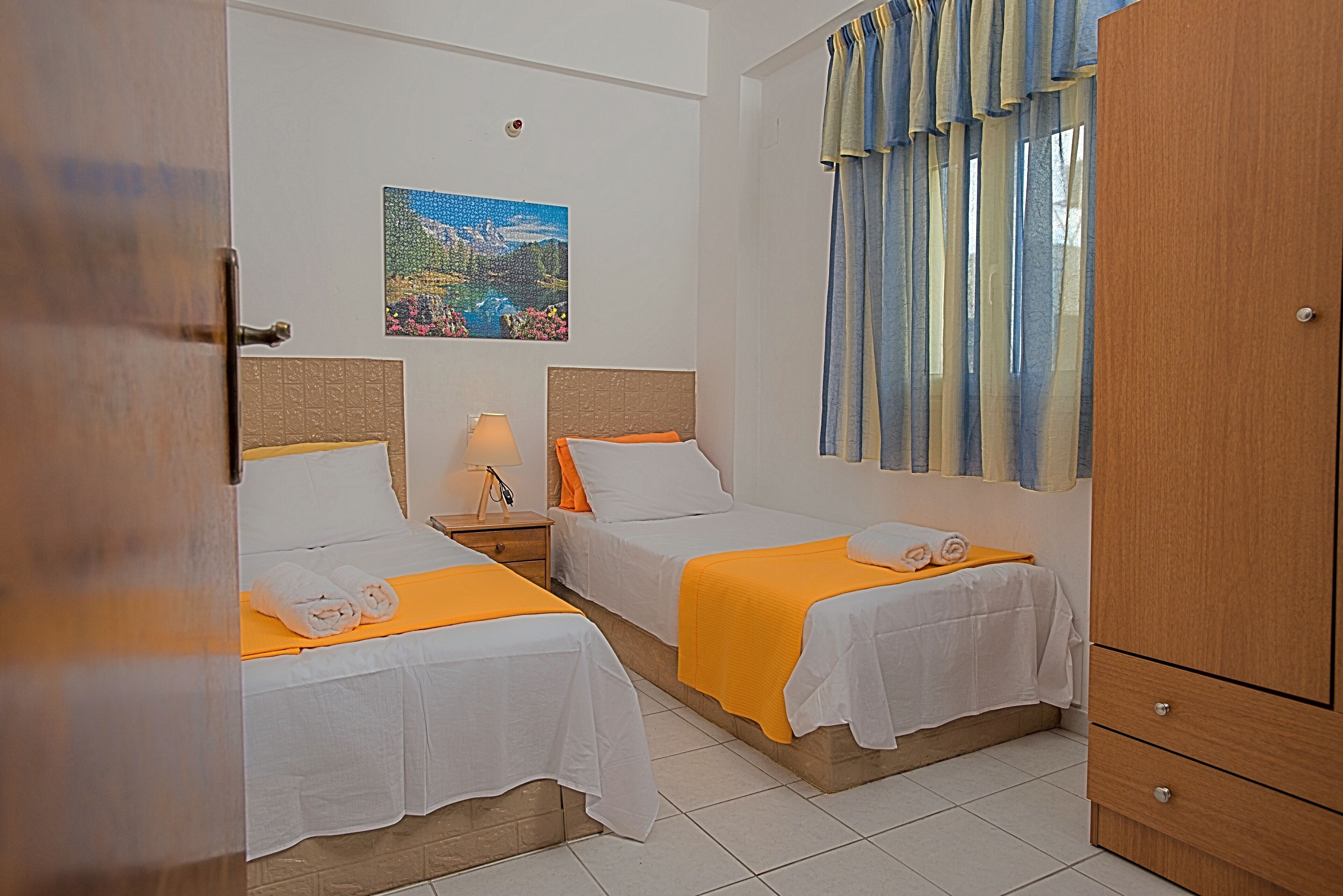 Une chambre d’hôtel avec deux lits, une table de chevet, une lampe, un tableau au mur et une fenêtre avec des rideaux.